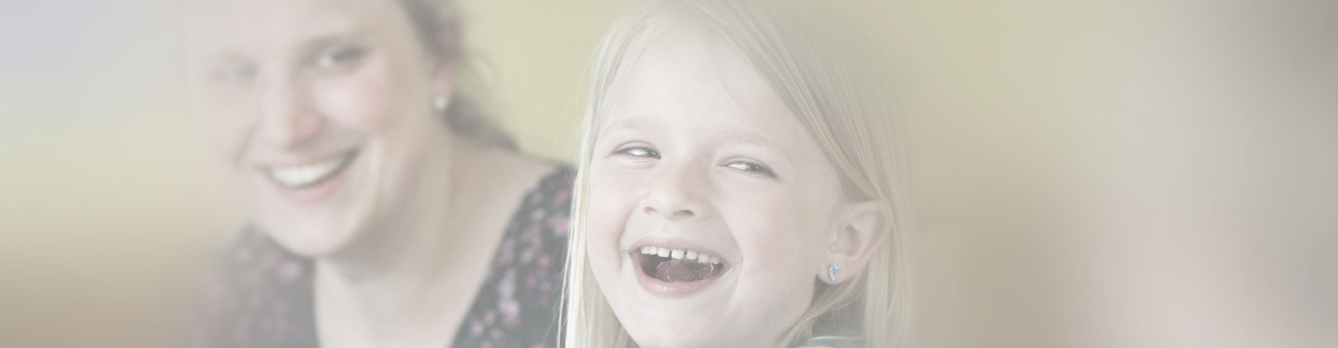 Foto eines lachenden Kindes mit zufrieden lächelnder Frau im Hintergrund.